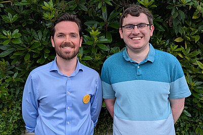 Adam Harley standing smiling on the left in a blue shirt and Grant Toghill on the right on a striped blue poloshirt, both with arms behind their back