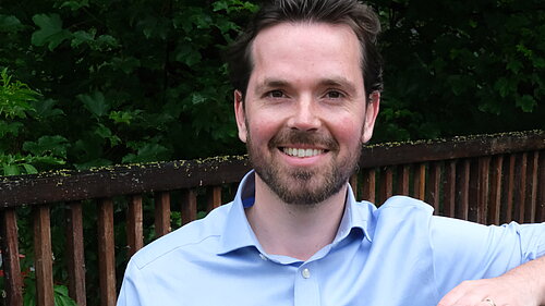 Adam Harley in a blue shirt standing with arm on top of a wooden fence of a bridge..
