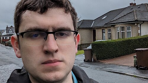 Grant Toghill, with serious looking expression, in a rain jacket with houses in the background.