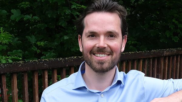 Adam Harley in a blue shirt standing with arm on top of a wooden fence of a bridge..