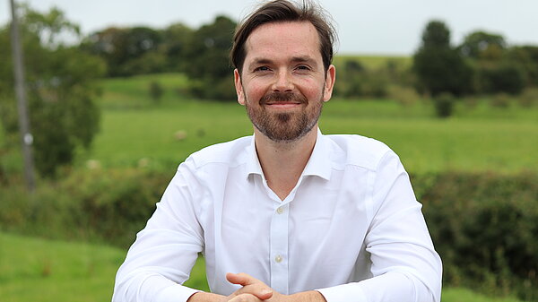 Adam Harley smiling while leaning on a fence with green fields in the background