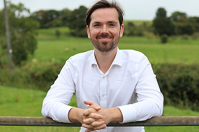 Adam Harley smiling while leaning on a fence with green fields in the background